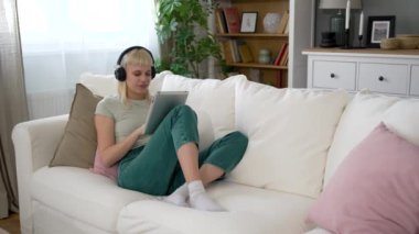 Genç yeniyetme kadın kanepeye oturmuş dijital tablet kullanıyor kablosuz kulaklıklarla müzik dinliyor internet araştırmasında çalışıyor, sosyal medyada tatil beldesi arıyor.