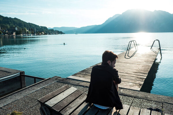Young man sitting on wooden bridge on pure lake surrounded with mountains, in Austrian Alps