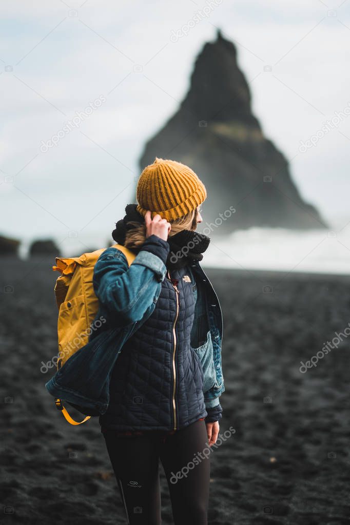 Young hipster woman with yellow backpack and hat on black beach of Vik i Myrdal, Iceland