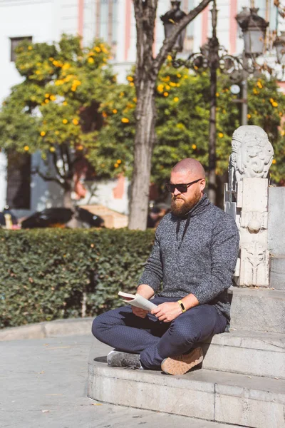Bearded man with shaved head relaxing by sitting and reading a book in Plaza del Triunfo (Seville, Spain)