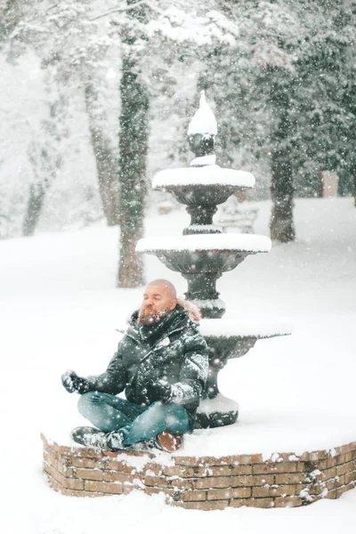 Man with beard and shaved head is sitting and meditating on the snow in a park or forest during a big snowfall - winter 2018 2019