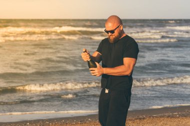 Kazılı kafa günbatımı ve gündoğumu tüm siyah arka plan üzerine bir deniz ile sahilde giyen sırasında bir açılan şampanya (köpüklü şarap) şişe sallayarak sakallı genç adam