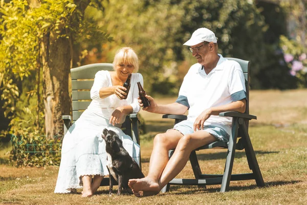 A retired couple is relaxing and having fun in the garden, drinking beer and playing with small dog