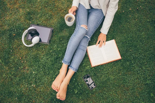 Top view of woman sitting in park (garden) on the green grass with smartphone, headphones, tablet, book and coffee in the hand