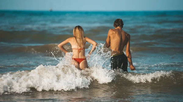 Young happy and sportive couple bathing in Adriatic sea in Rimini (Italy) - sporty man and woman go into rough water (sea or ocean) with waves