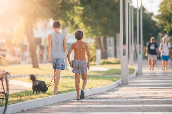 Two young guys (boys) best friends or brothers are walking with a little black dog on summer Italian street during sunset or sunrise - travel, vacation in Italy on seaside and friendship concept