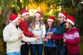 Skupina šesti nejlepších přátel s zimní svetry a červený santa klobouky, slavit Vánoce s šampaňským a ohňostroj (prskavky) na západ slunce v horké středomořské nebo Rovníkové země