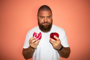 Young man with white t-shirt and big beard making difficult choice between red apple and pink donut (already bitten). Against orange background. clipart