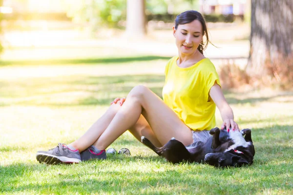 草の上に座っていると 日当たりの良いガーデンで楽しいカジュアルな夏服の女の子の画像 オランダ オランダの都市公園で小さな黒い犬と遊ぶ若いブルネットの幸せな女 — ストック写真