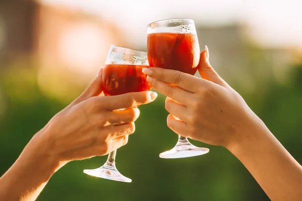 Cheers. Female hands cheering on sunset with red wine fresh sangria glasses with green sunny garden background - female friendship, holidays celebrating and party concept
