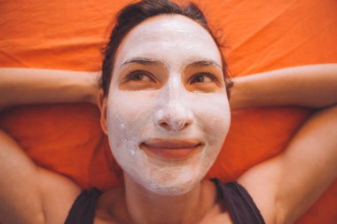 Portrait of happy young beautiful smiling sporty woman with white facial clay mask relaxing on the bed - Beauty treatments for face, skin and body care, healthy lifestyle and love yourself concept clipart