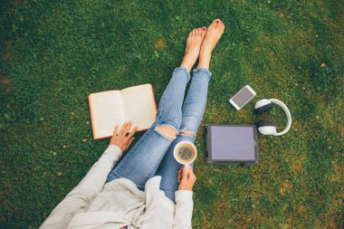 Park (Bahçe) smartphone, kulaklık, tablet, kitap ve kahve el ile yeşil çimenlerin üzerinde oturan kadının üstten görünüm
