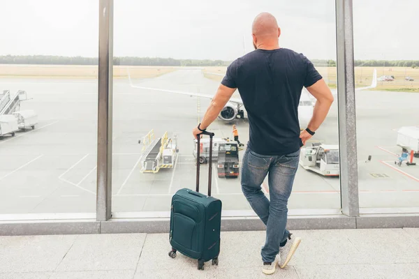 A man in the airport is watching preparation of an airplane while is waiting for his flight, view from behind