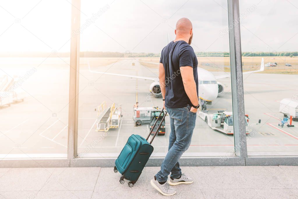 Sporty man with shaved head and beard is observing an airplane preparation while waiting his flight. Sunny warm shot.