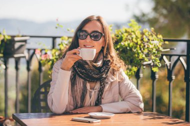 Çekici kız bir barda oturan bahar veya sonbahar/kış aylarında güneşli bir gün dağ manzaralı tablo. O gülümseyen ve ile onun smartphone masada bir fincan kahve (kapuçino) tutan.