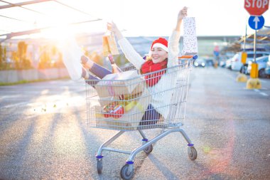 Mutlu genç gülümseyen kadın oturan bir sürü hediye kutuları ve kırmızı Santa Noel giyen onun elinde iki alışveriş torbaları ile alışveriş sepetindeki şapka - tatil satın alma ve kutlama kavramı.