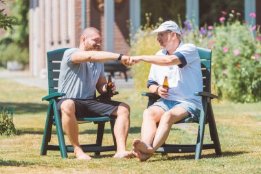 Two men drinking beer together and making power five gesture in summer sunny garden (Netherlands - Holland) - male friendship and family party concept clipart