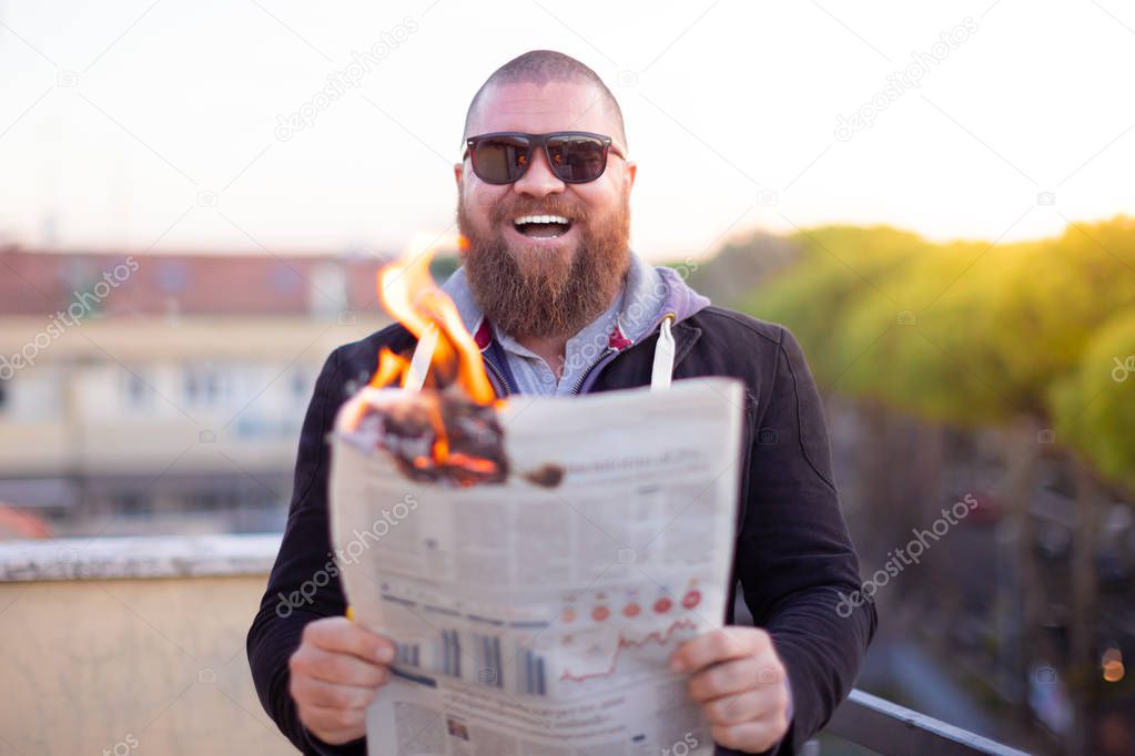 Portrait of excited (happy) bearded man looking to the newspaper (on fire) - burning magazine in man's hands - hot and breaking news concept - laughing about financial crisis
