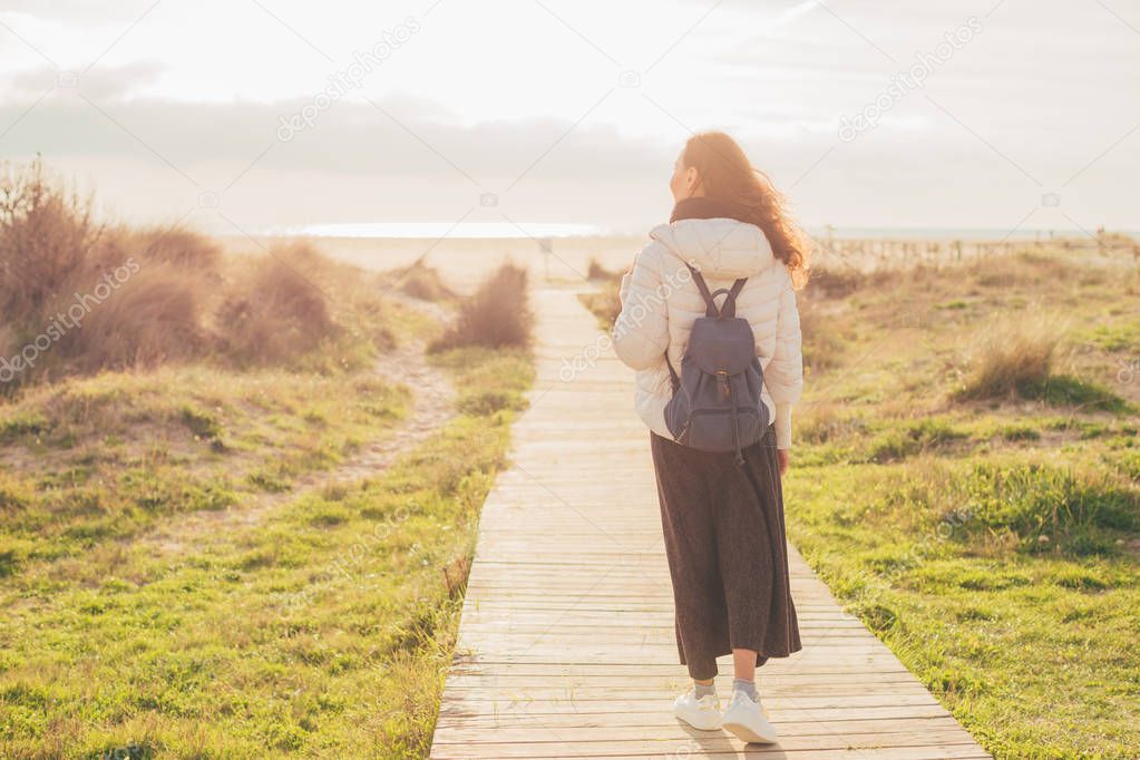 Rear view of young beautiful romantic brunette woman walking on boardwalk looking at the ocean. Peace of mind, freedom and travel concept. Tarifa, Spain.