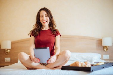 Genç kadın yatakta kahvaltı ve elleri tablet ile otel odasında oturuyordu