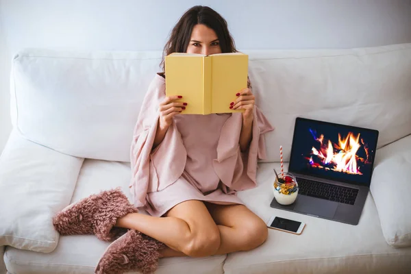 Girl hiding her face behind a book while sitting on the sofa with a fireplace on the laptop - young woman relaxing and reading in cozy room - hardcover book mockup