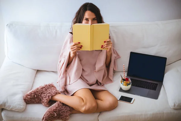 Girl hiding her face behind a book while sitting on the sofa with a laptop and yogurt or ice cream with fresh fruits in a jar - young woman relaxing and reading in cozy room - hardcover book mockup