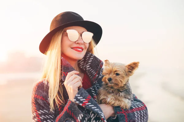 美しい笑顔のヒップスター トレンディな金髪女性のサングラス 黒い帽子を身に着けている小さな犬とビーチを歩いて肖像画 — ストック写真