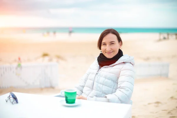 Portrait of a young smiling woman sitting at the table with a cup of coffee on a winter ocean beach cafe during a beautiful sunset or sunrise on the background, Tarifa (Spain)