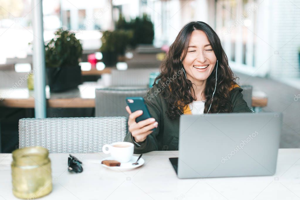 Happy asian smiling millennial girl looking at the screen of laptop, holding a smartphone in her hands - working in a cafe while traveling, digital nomading, watching movie, video calling concept