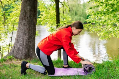 Kırmızı ceketli genç kız yazın doğada yoga minderini açıyor.