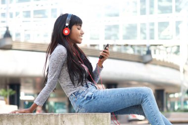 Akıllı telefon ve kulaklık ile müzik dinlemek şehir dışında oturan gülümseyen Genç Hintli kadın portresi yan