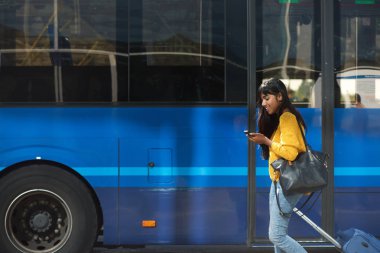 Yan çanta ve cep telefonu ile otobüs istasyonu tarafından yürüme gülümseyen Genç Hintli kadın portresi