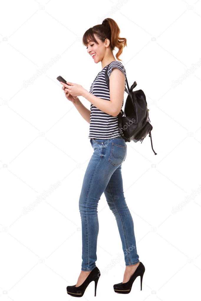 Full length side portrait of young woman walking with mobile phone and bag against white background