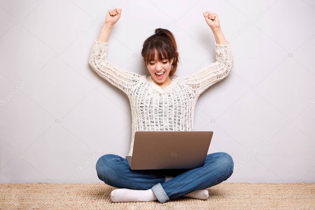 Portrait of a cheerful young woman sitting on floor with laptop and arms raised