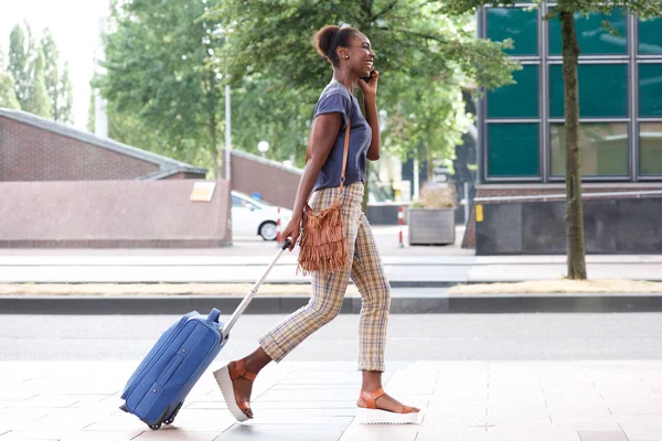 携帯電話とスーツケースが付いている都市を歩いて幸せなアフリカ系アメリカ人女性の完全なボディ側の肖像画 — ストック写真