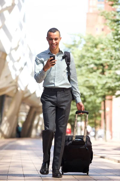 Full body portrait of traveling businessman walking in city with suitcase and mobile phone