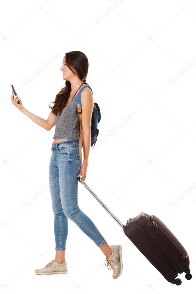 Full length side portrait of smiling asian woman walking against isolated white background with suitcase and cellphone