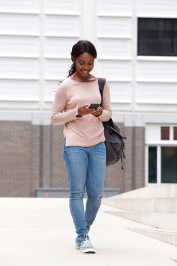 Şehir içinde cep telefonu ile yürüyen gülümseyen genç Afro-Amerikan kadın portresi tam uzunlukta