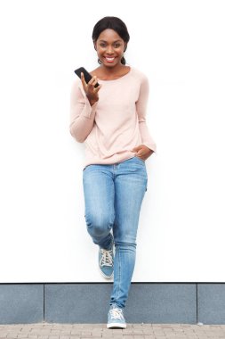 Cep telefonu ile beyaz duvar karşı duran gülümseyen genç siyah kadın portresi tam uzunlukta