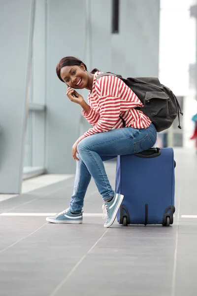 駅でスーツケースの上に座って 携帯電話で話し陽気の若いのアフリカ系アメリカ人の女性の肖像画 — ストック写真