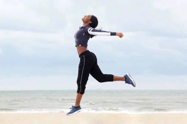 Full body side portrait of african american woman running and jumping with arms raised at beach