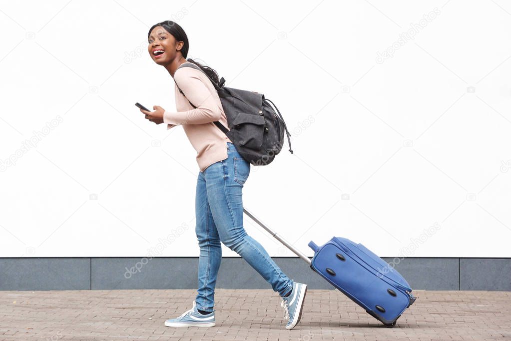 Full length profile portrait of young female traveler walking with cellphone and suitcase against white wall