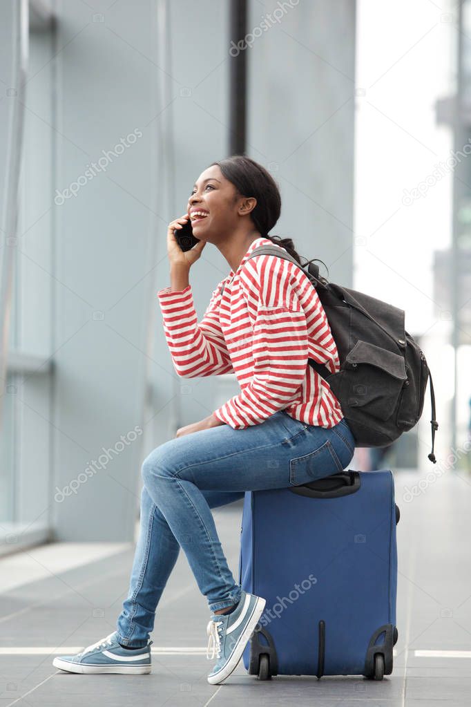 Portrait of happy young african american woman sitting on suitcase and talking with mobile phone at the station 