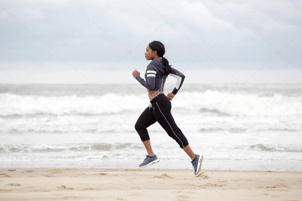 Full length side portrait of young african american woman running on the beach by the water