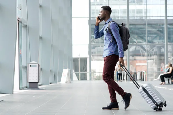 スーツケースと空港で携帯電話で旅行する若いアフリカ人の完全なボディ側の肖像画 — ストック写真