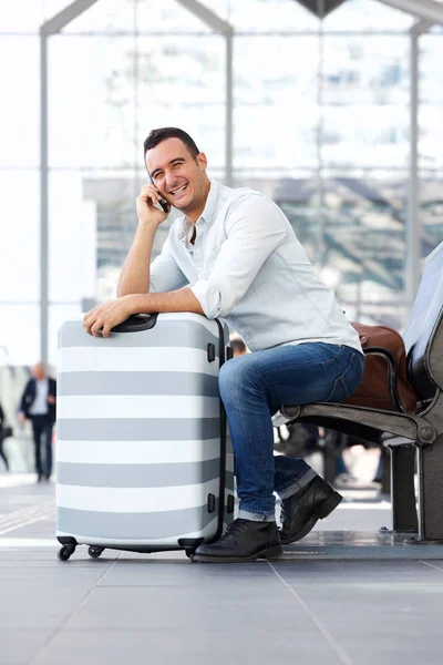 スーツケースを持って座っている ターミナル空港で携帯電話で話している幸せな男の肖像 — ストック写真