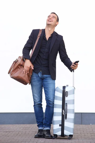 Full length portrait of happy travel man by white wall with suitcase bag and mobile phone