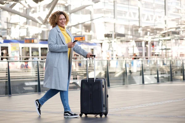 Full body side portrait of happy travel woman walking in station with suitcase and mobile phone