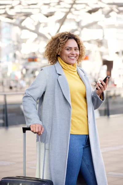 Portrait of smiling african american woman with suitcase and mobile phone at station