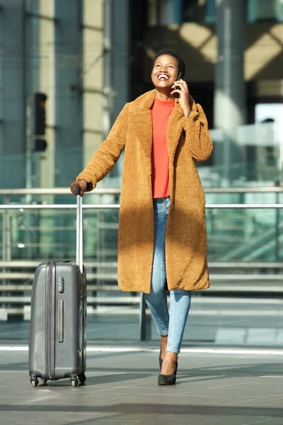 Full body portrait of happy young black woman walking in station with suitcase and mobile phone